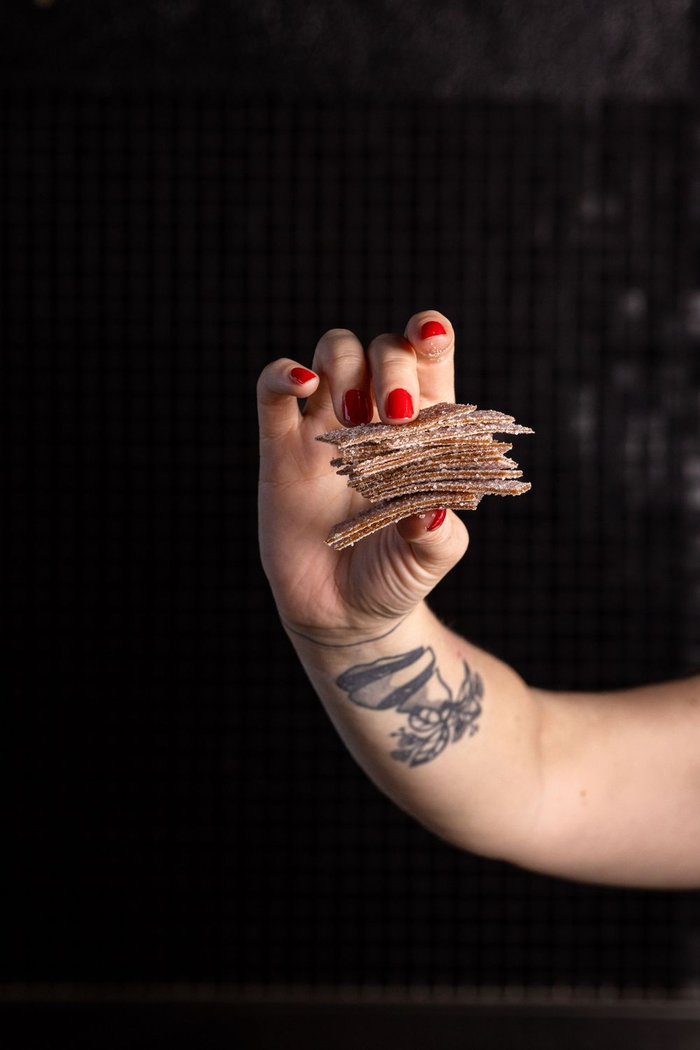 A tattooed arm with red painted nails holds a piece of torn brown cardboard against a dark background.