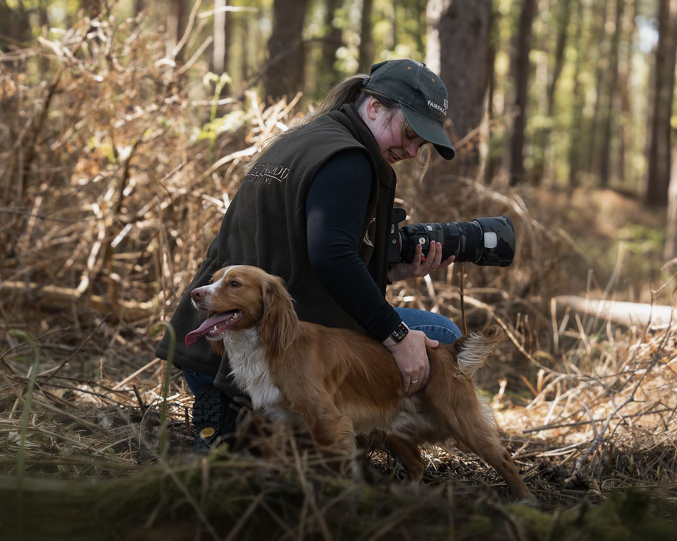 A photographer crouches beside a cocker spaniel in a sunlit woodland setting.