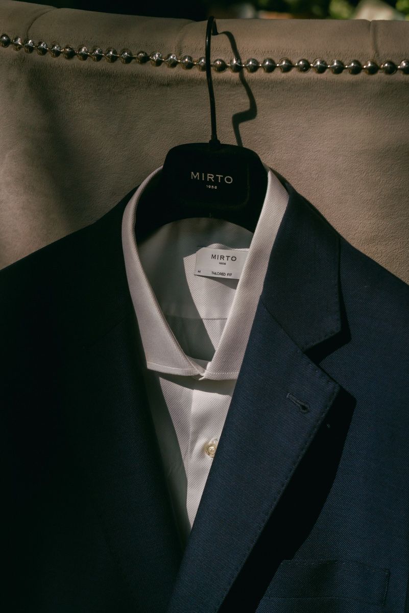 Navy blue suit jacket with white dress shirt on a hanger against dark background.