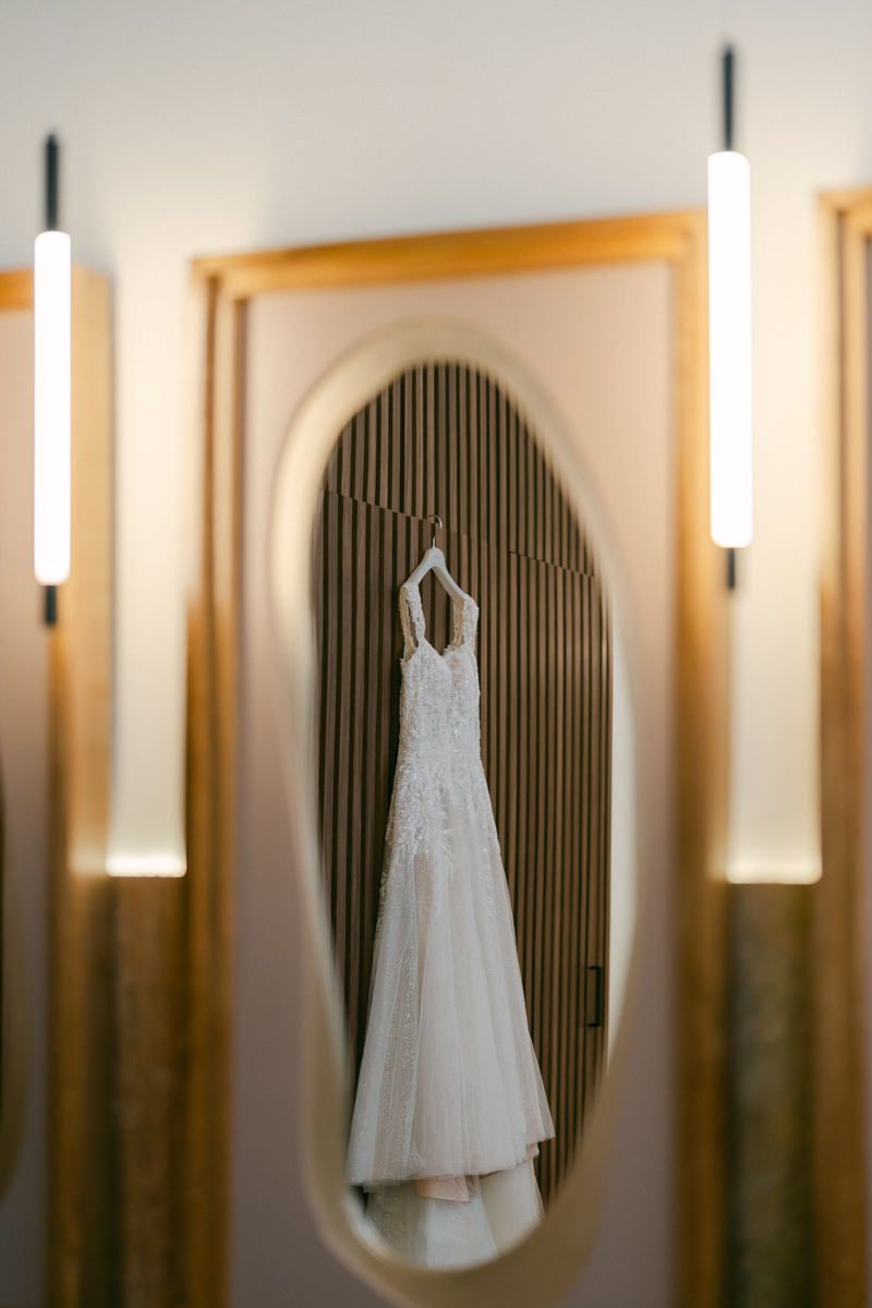 White wedding dress hangs elegantly against dark wall with oval mirrors and vertical lighting fixtures in background.