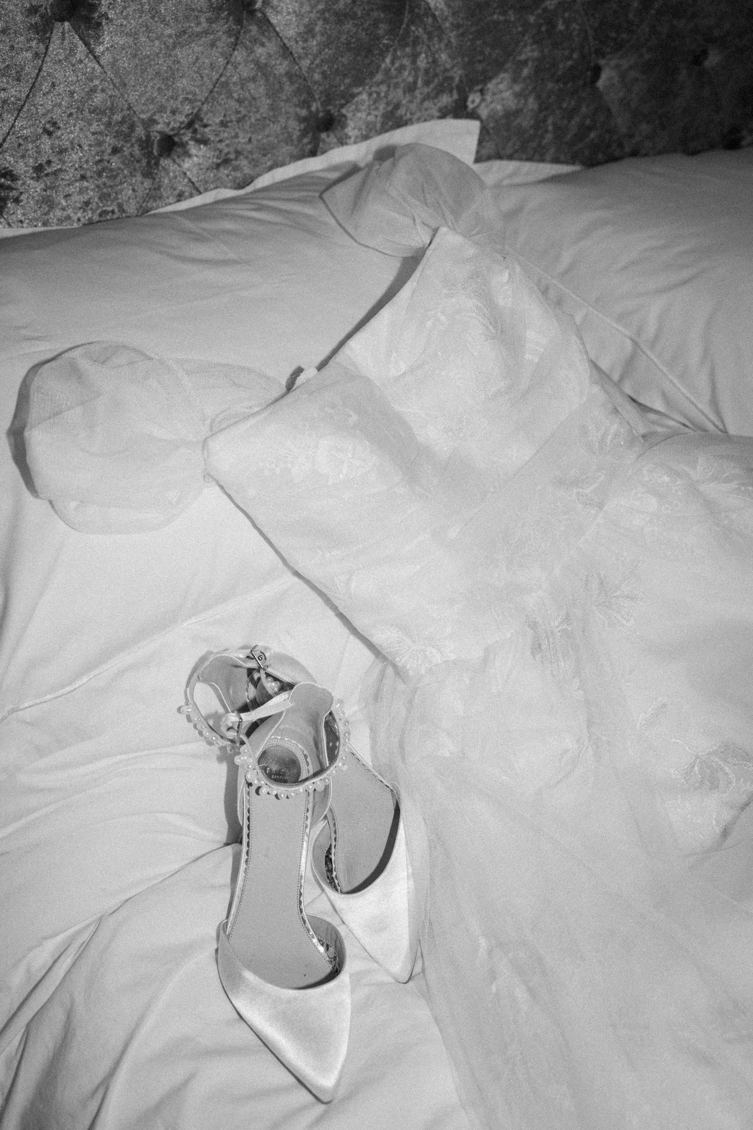 Black and white photo of a white satin bridal heeled shoe placed on crumpled bedding.