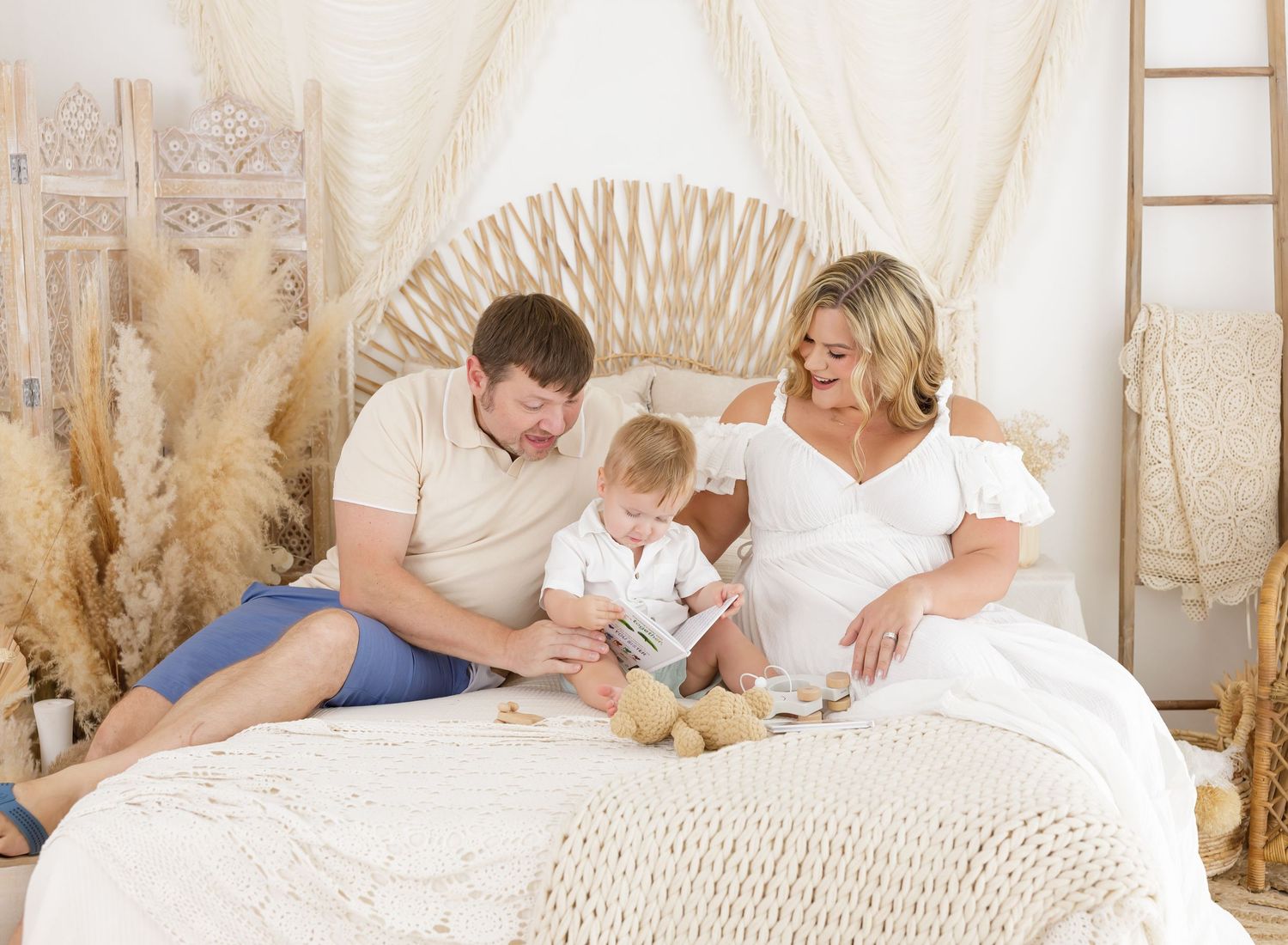 Family relaxes together on white bed with boho decor and pampas grass in background.
