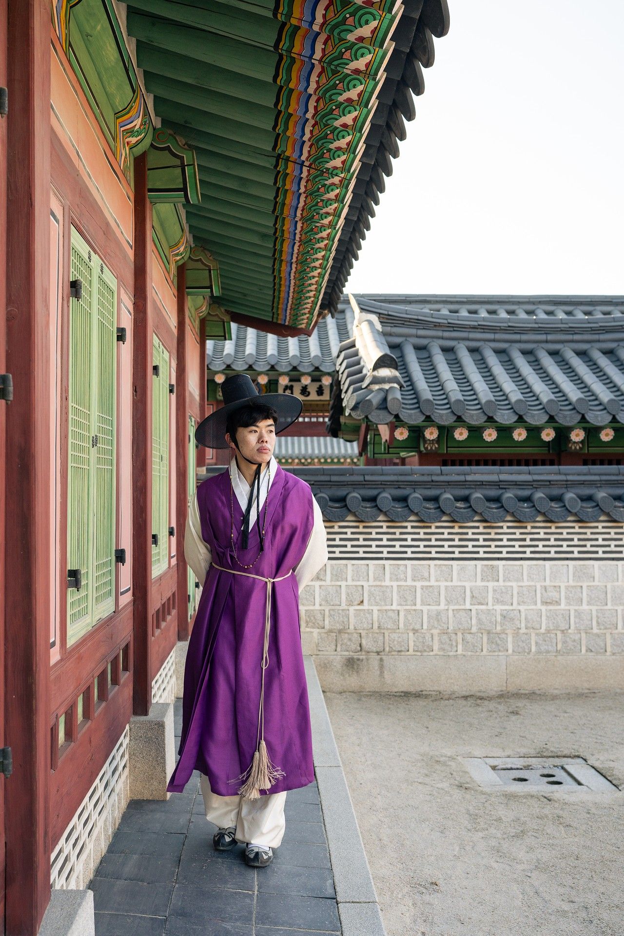 People in traditional Korean hanbok attire walk along a covered walkway at a historic palace complex.