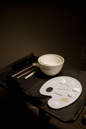A white ceramic mixing bowl and artist's palette sit on a dark surface in moody lighting.