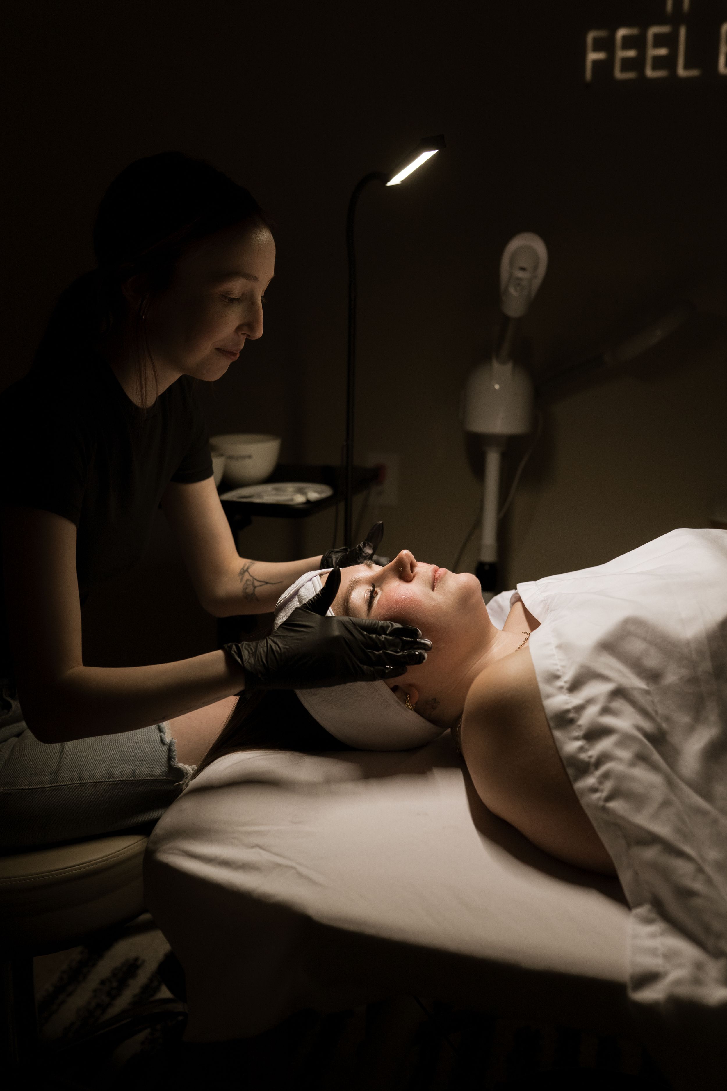 A beauty therapist performs a facial treatment on a client in a dimly lit spa room with soft lighting.