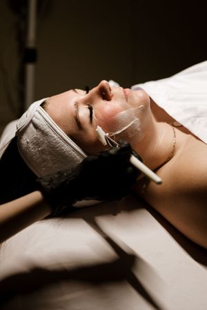 Someone receiving a relaxing facial treatment at a spa with a white face mask and headband while lying on treatment bed.