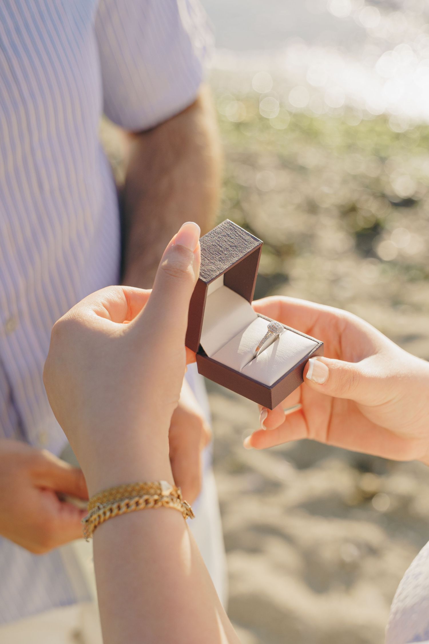 Close-up sequence of hands opening a small jewelry box revealing a gold chain bracelet on a sunny beach day.