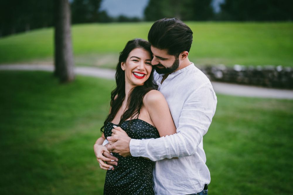 Couple sharing a playful embrace on a lush green lawn with pine trees.