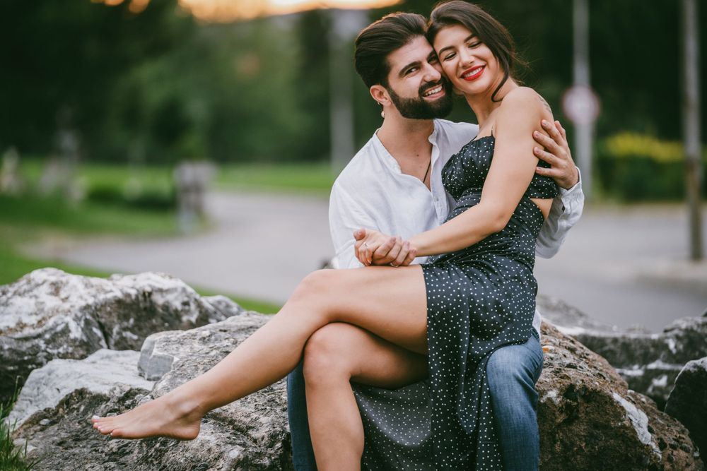 Joyful couple laughing together while sitting on large rocks at dusk.