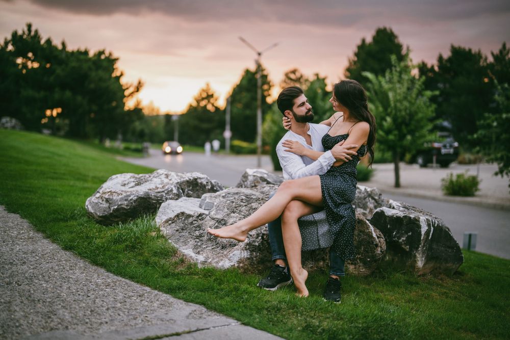 Romantic couple embraces on large rocks in park at sunset with green lawn and trees in background.