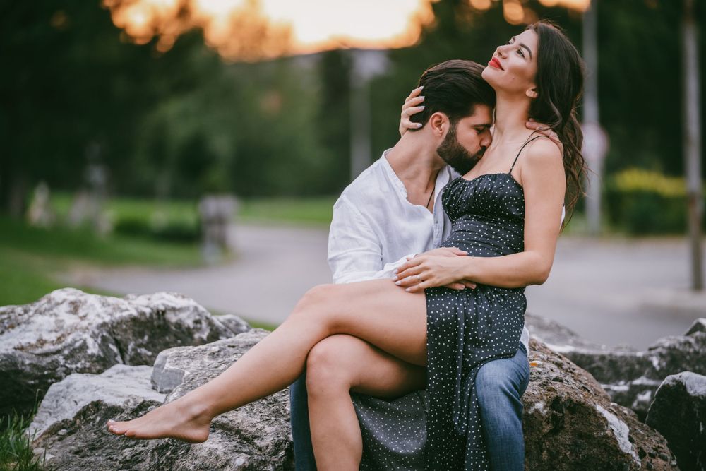 Tender moment between couple sitting on natural stone formation in park setting.