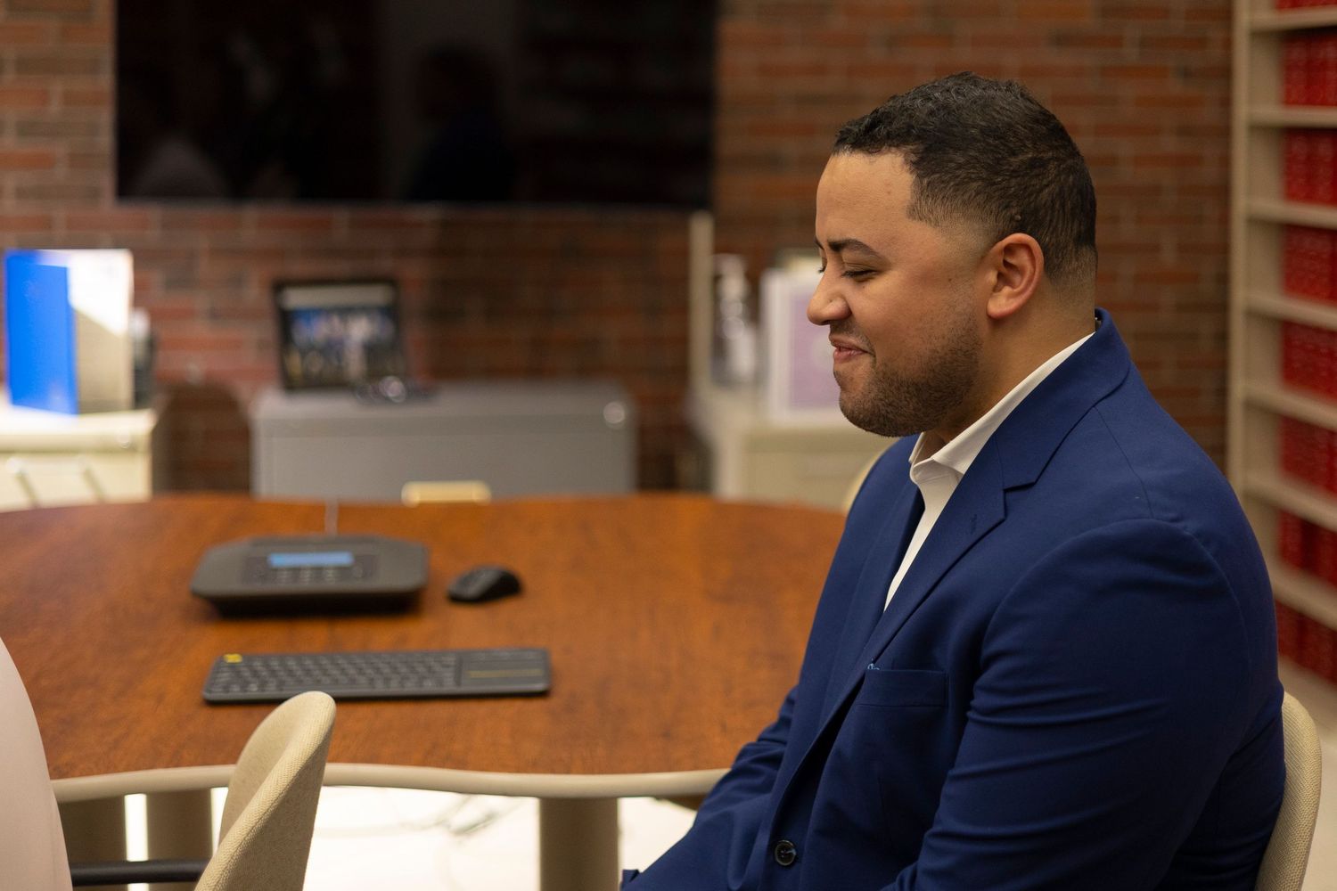 Professional in a navy blue suit sits at a desk in an office setting with brick walls and computer equipment visible.