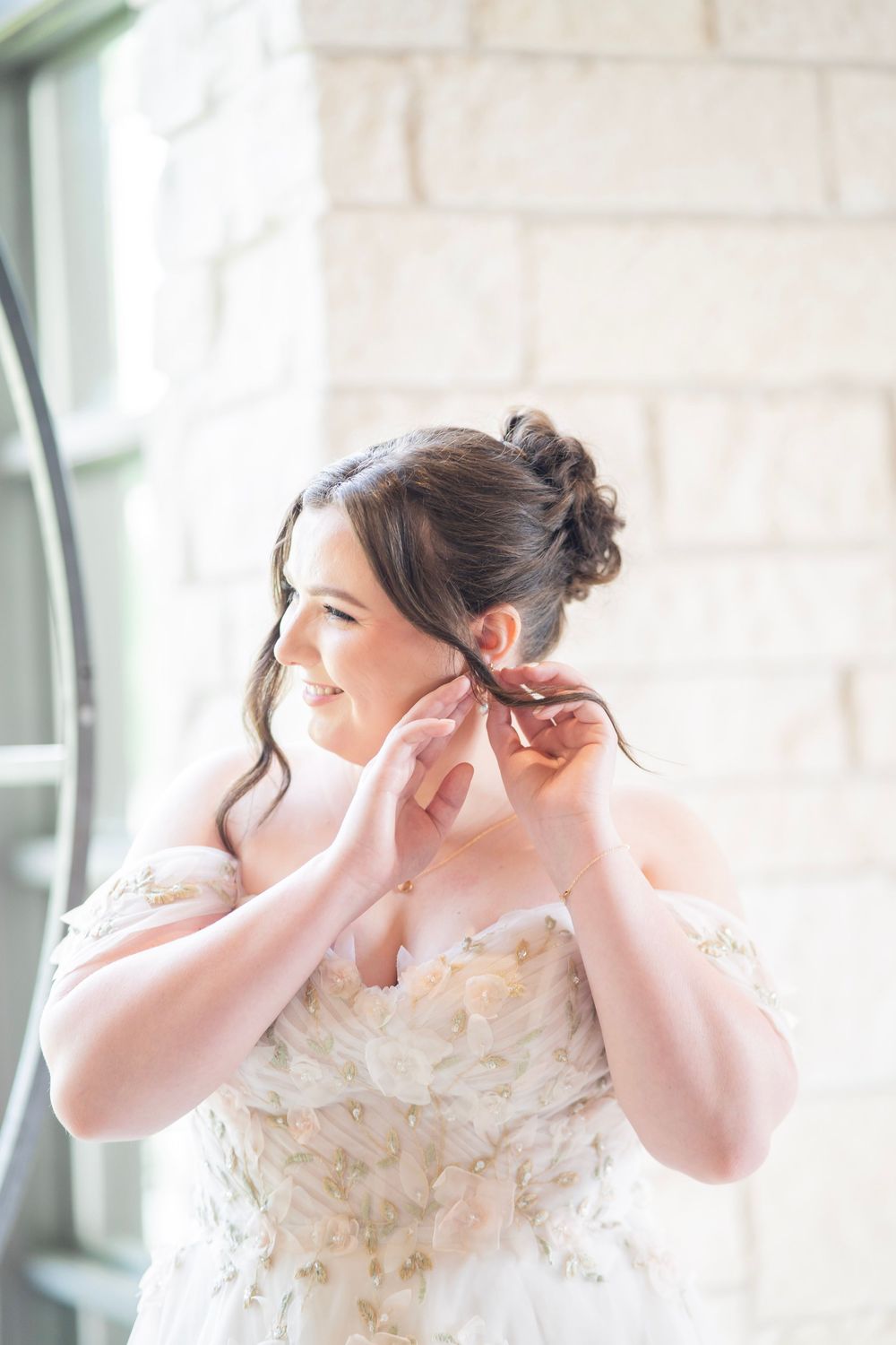 Bride in off-shoulder floral wedding gown putting on earrings near stone wall window.