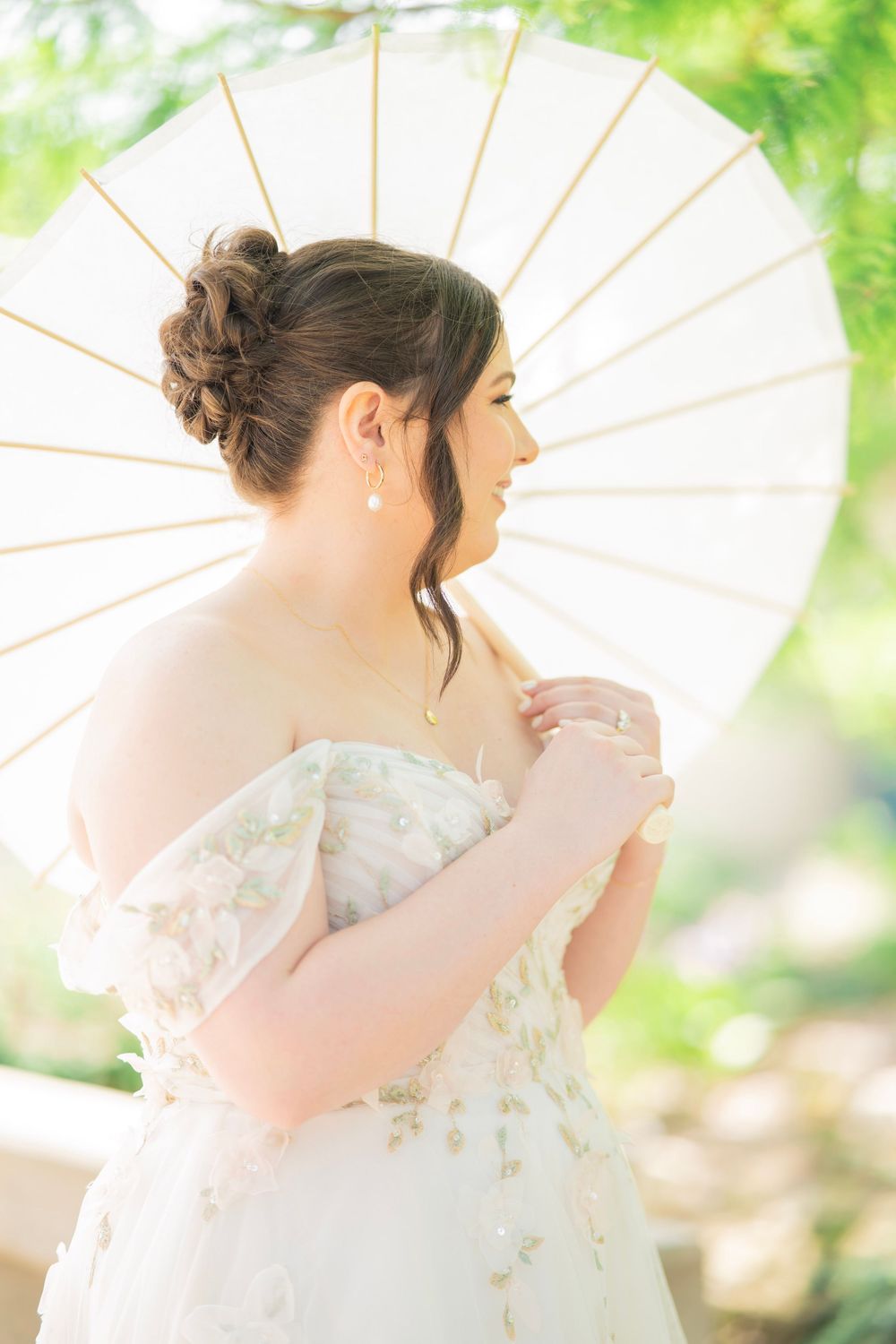 Bride in floral off-shoulder wedding gown holding white parasol with elegant updo hairstyle outdoors.