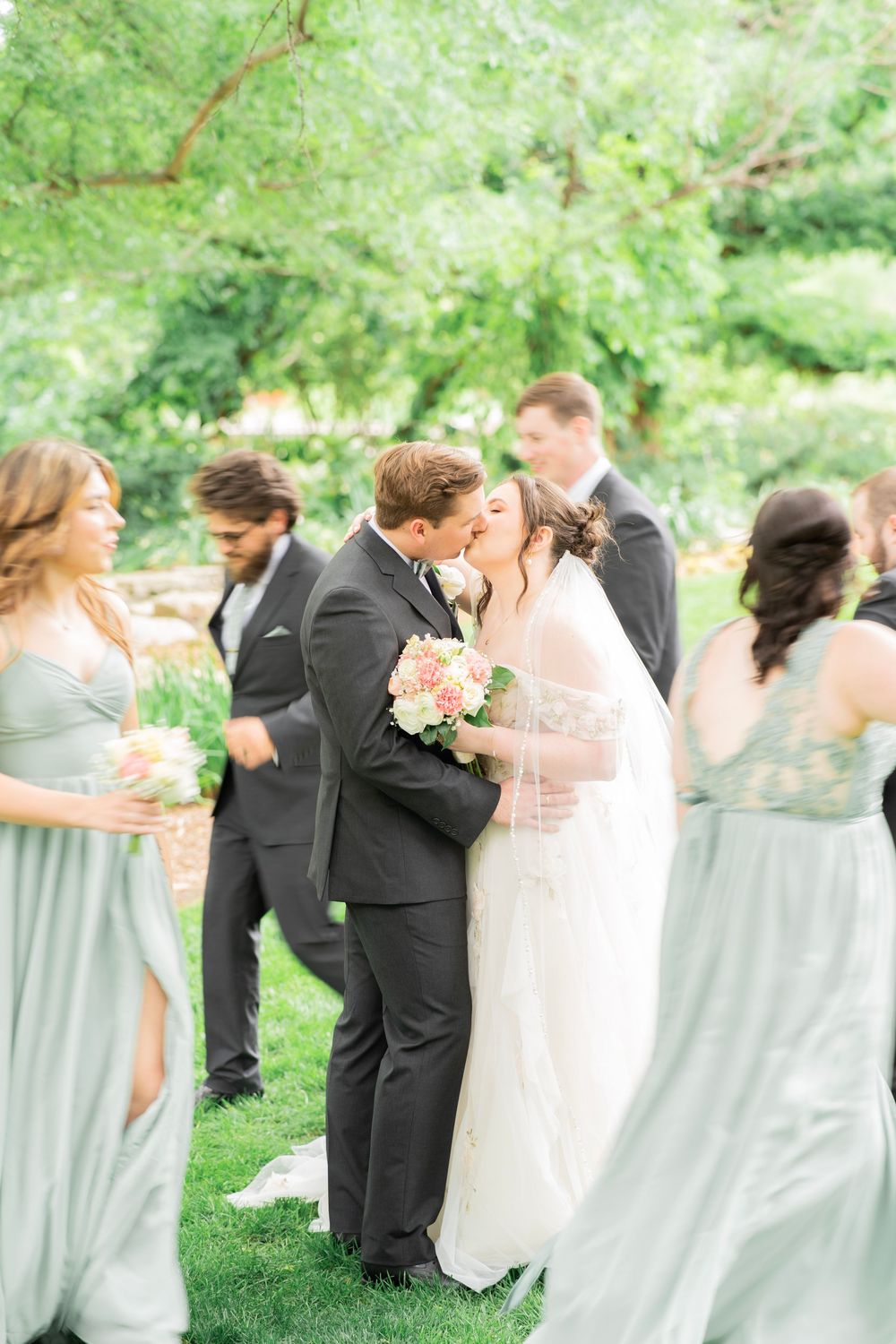 Bride and groom share a romantic kiss surrounded by wedding party in a sunlit garden setting.