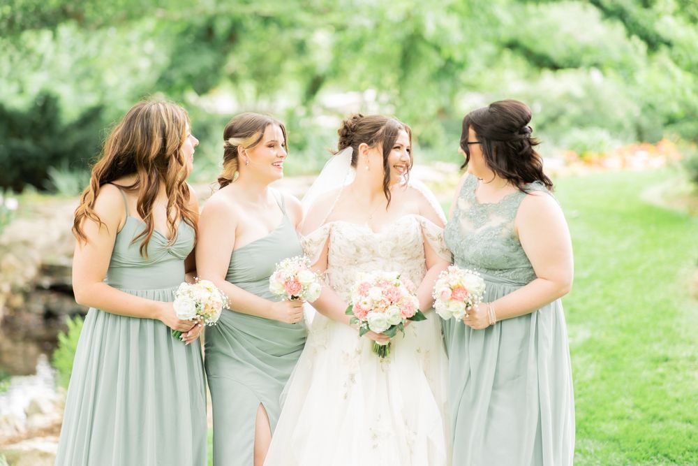 Bride with three bridesmaids in sage green gowns walk together smiling, holding pink and white bouquets outdoors.