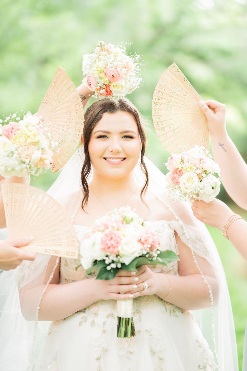 Smiling bride in lace gown holds pink bouquet surrounded by bridesmaids holding decorative fans in sunlit garden.