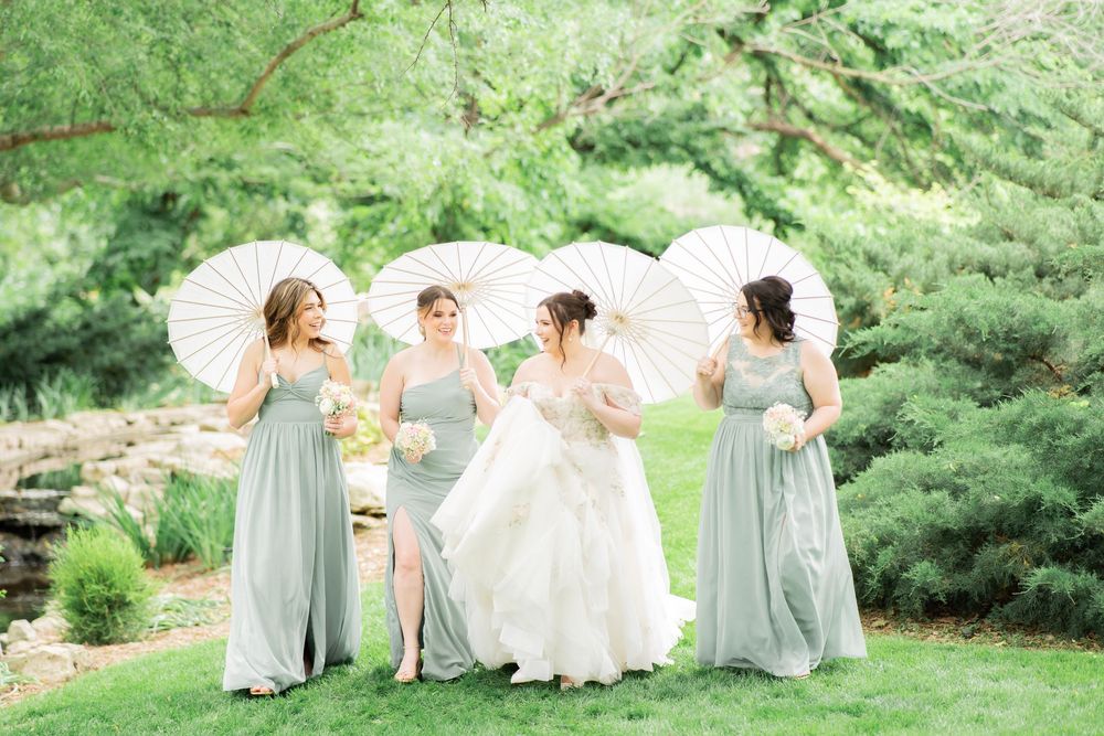 Bride and three bridesmaids in sage green dresses pose with white parasols in a lush garden setting.