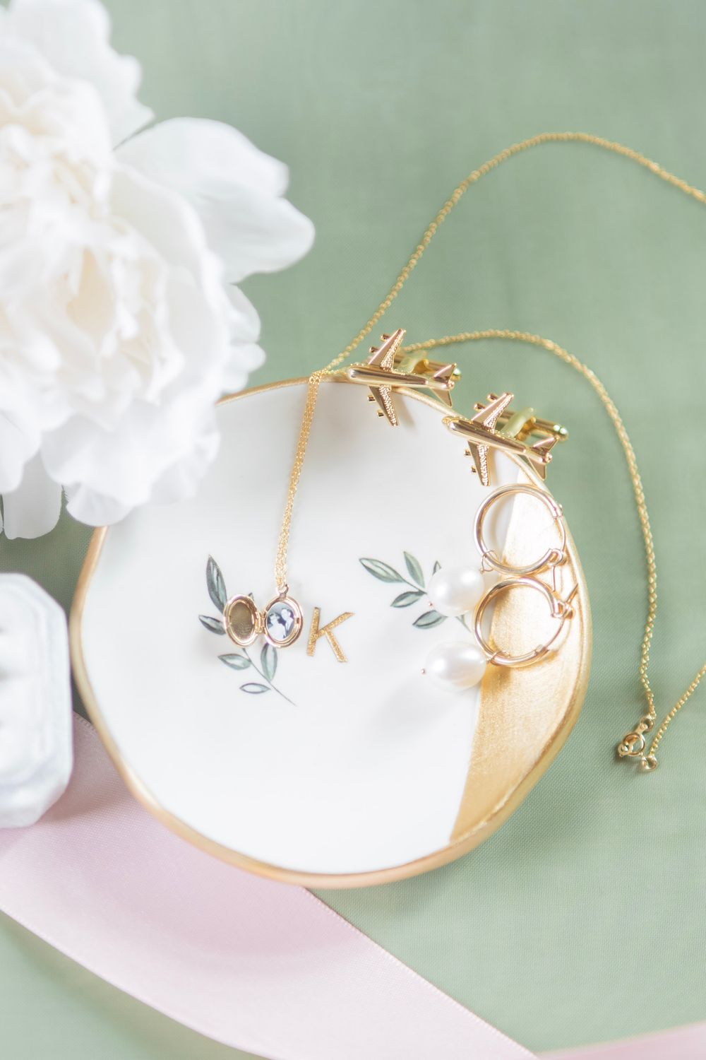 Gold and white bridal jewelry dish with rings, necklace, and arrow earrings on sage green background.