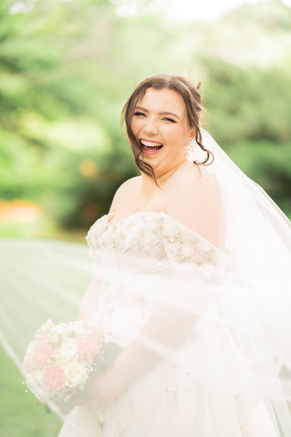 Laughing bride in lace wedding dress holding pink and white bouquet with veil flowing.