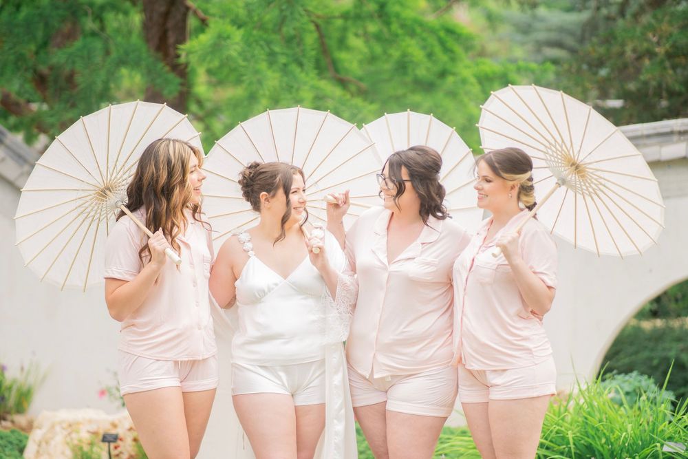 Bride and four bridesmaids in pink pajamas holding white parasols outdoors in garden setting.