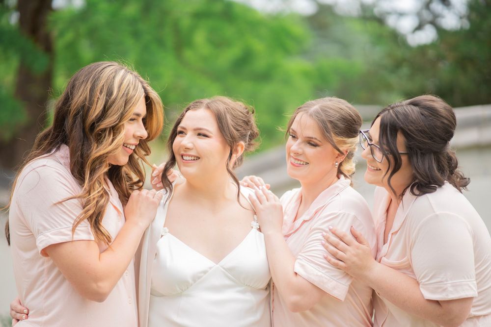 Bride in white gown laughing joyfully with three bridesmaids wearing blush pink tops outdoors.
