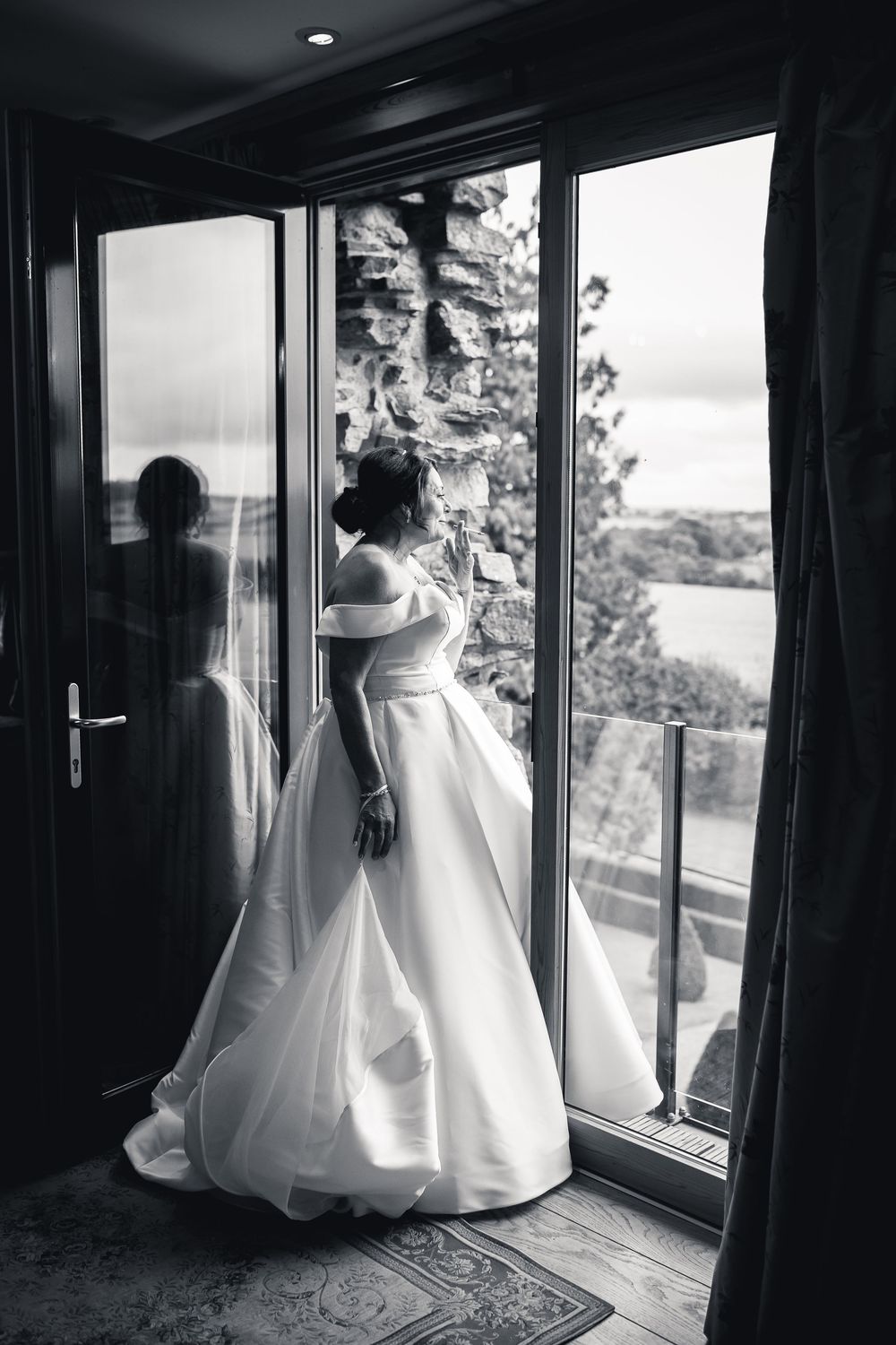 A bride in an elegant white wedding gown gazes out through glass doors onto a balcony in black and white photography.