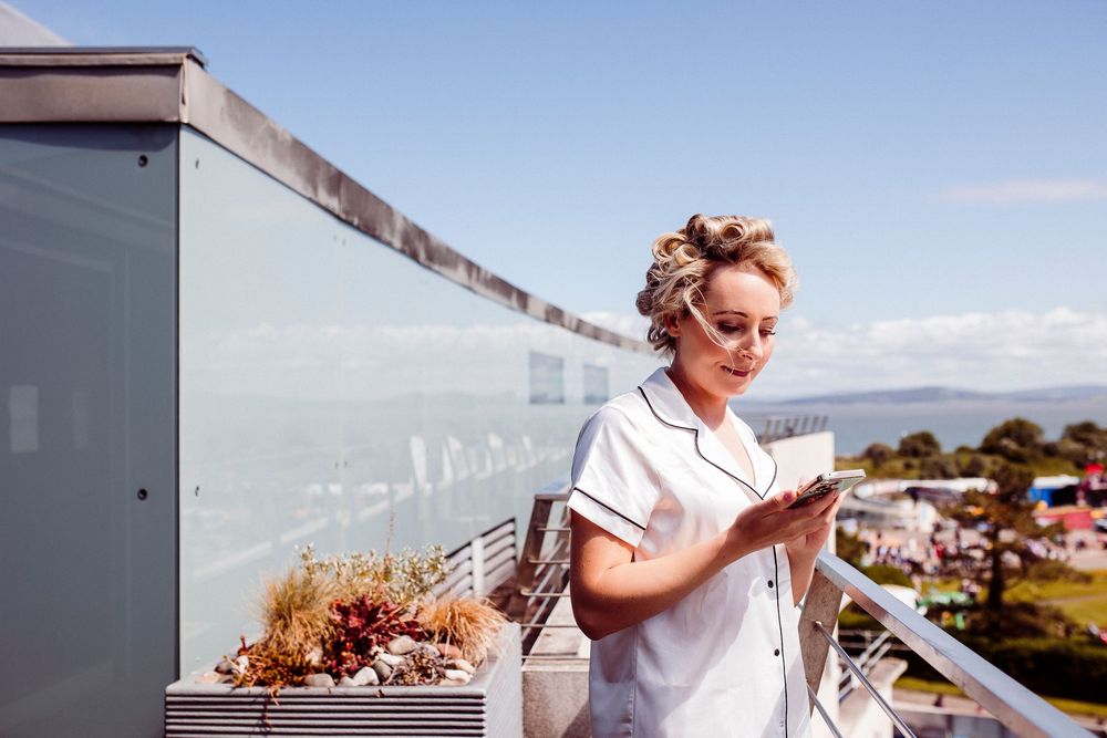 Person in white t-shirt checking phone on sunny balcony with ocean view in background.