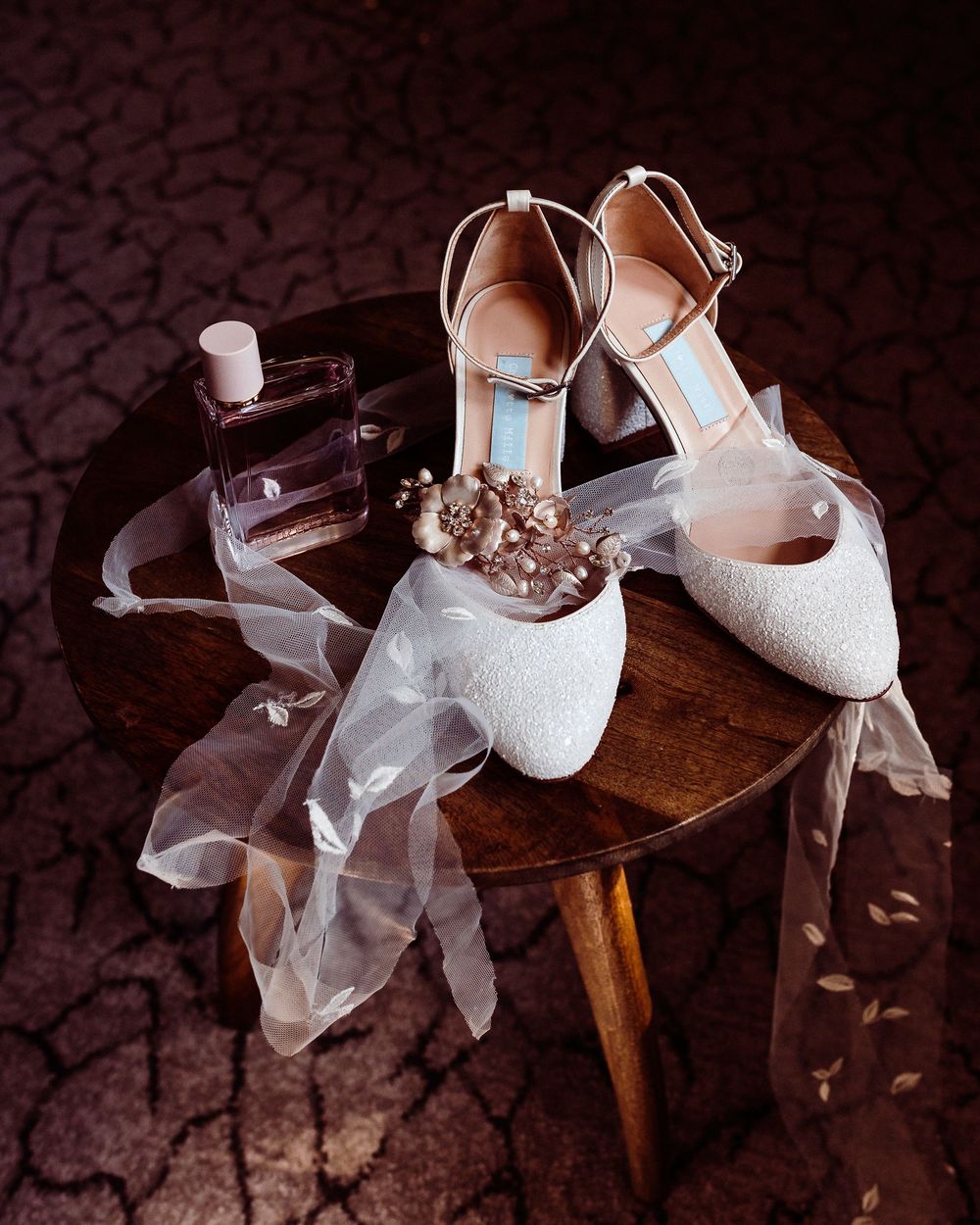 Elegant white bridal shoes with delicate lace details and sheer tulle veil displayed on dark wooden surface.