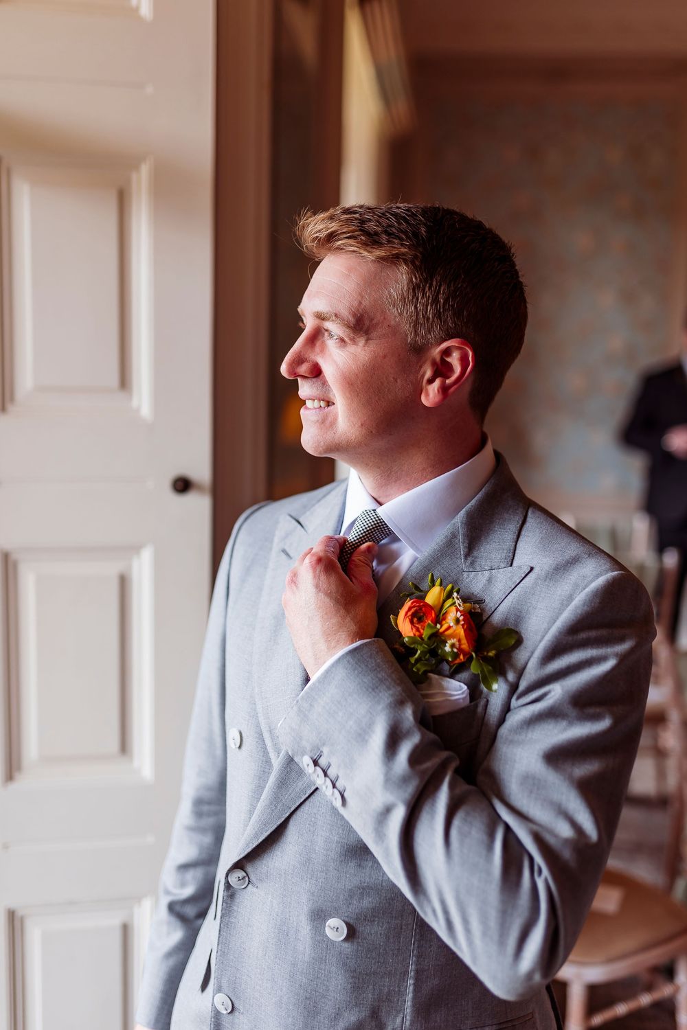 A groom in a light gray suit with an orange boutonniere looks out a window in a series of candid shots.