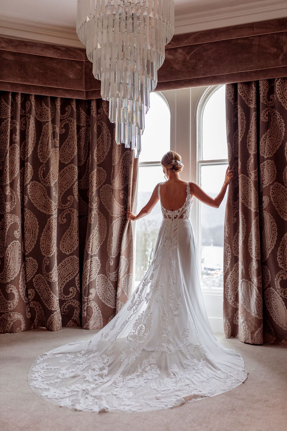 A stunning white lace wedding dress with a long cathedral train elegantly displayed against rich brown curtains.