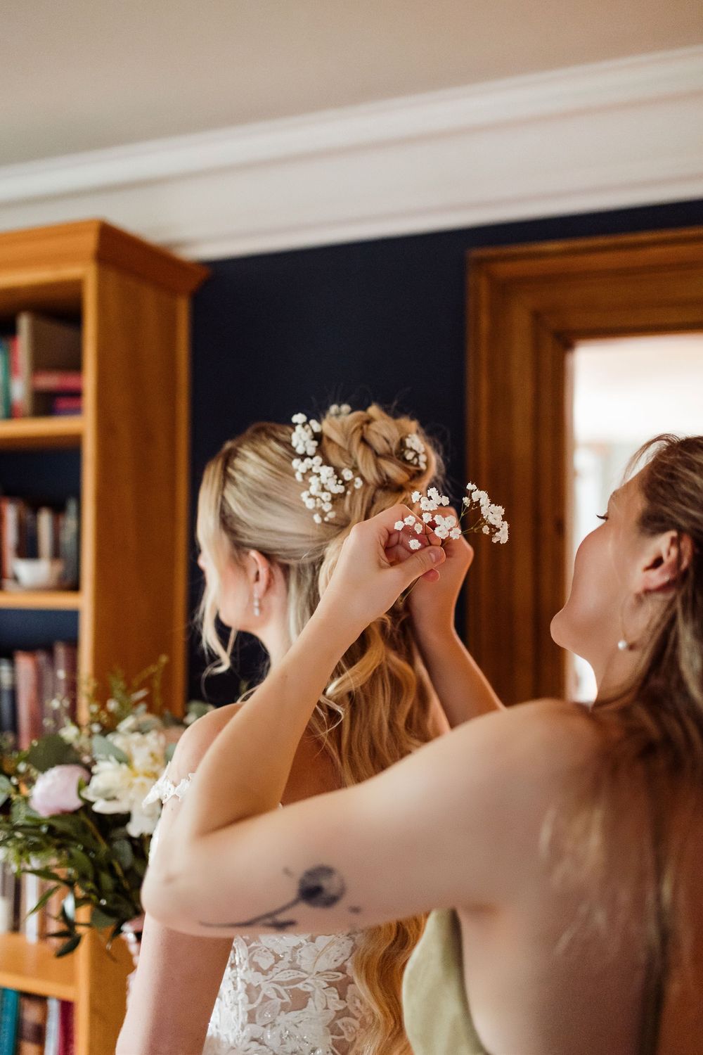Wedding preparations show delicate white flowers being placed in an updo hairstyle with a wooden bookshelf in background.