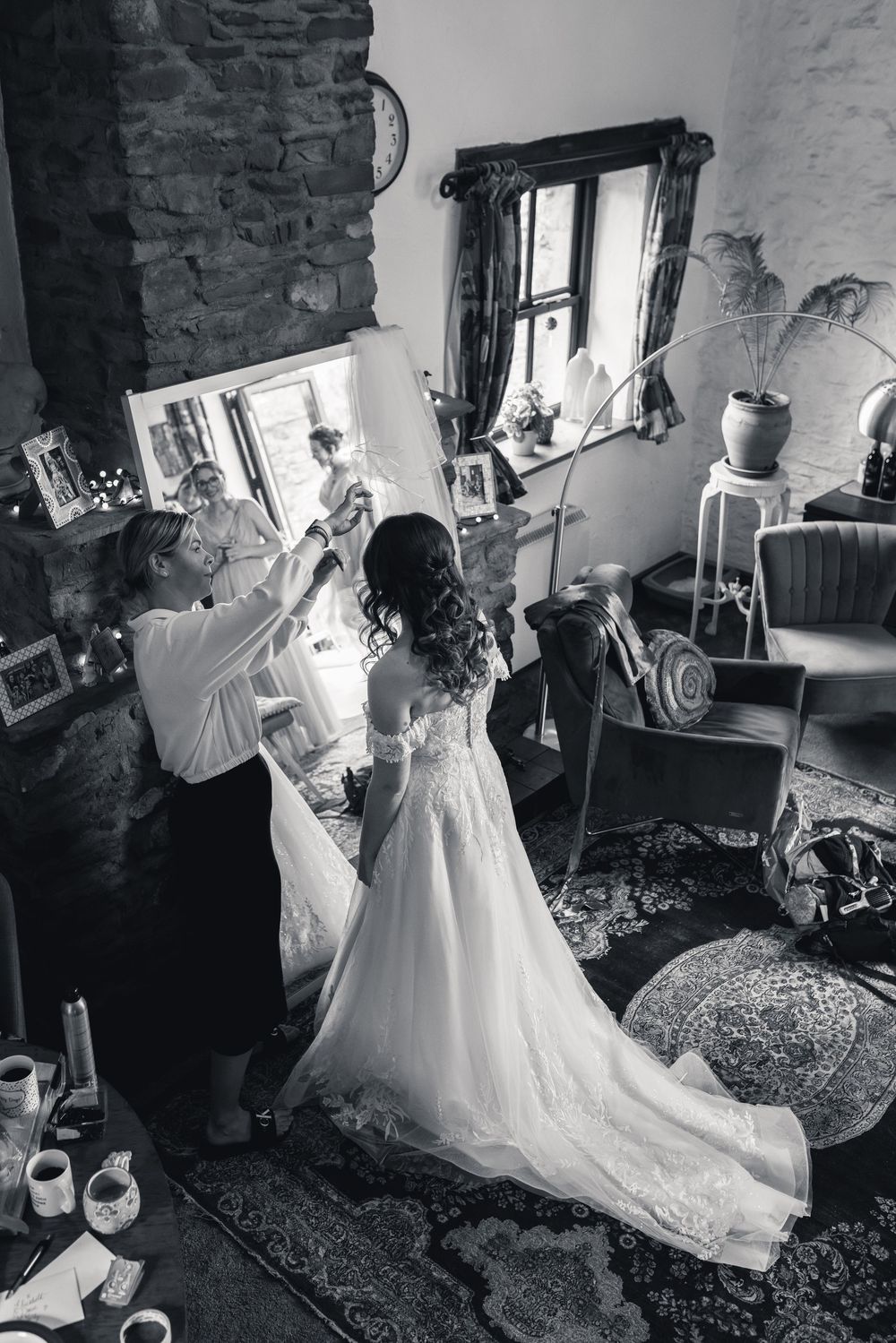 Black and white photo of wedding dress preparations in a rustic room with natural light streaming through windows.