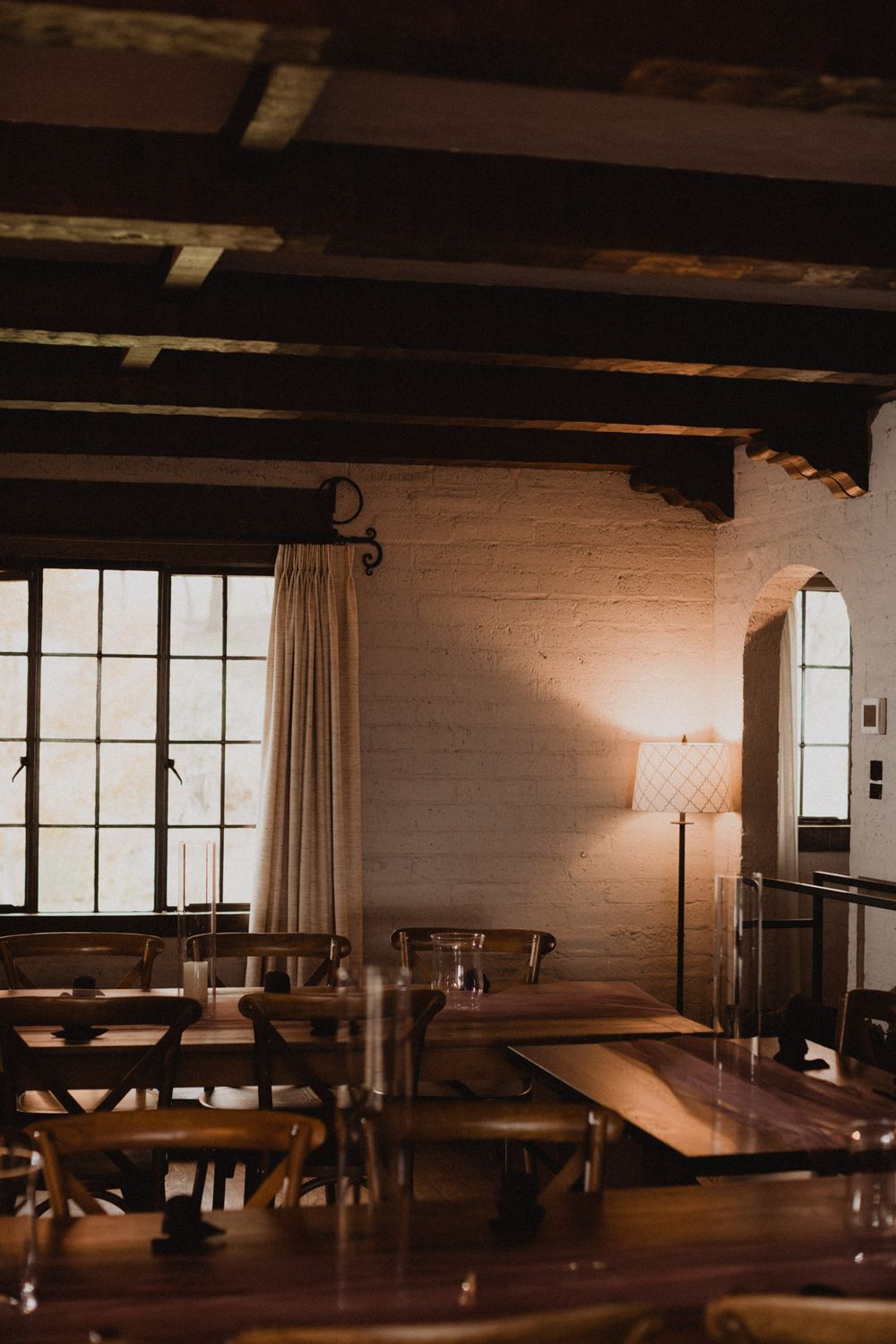 Rustic restaurant interior with wooden tables and exposed beams illuminated by warm sunlight through large windows.
