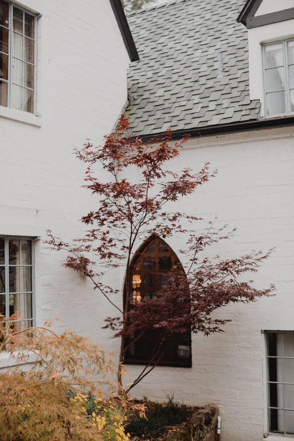White painted brick house exterior with Japanese maple trees and gray shingled roof against cloudy sky.