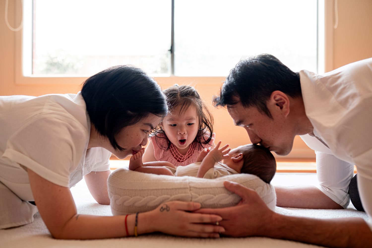 A tender Melbourne family moment with parents kissing their newborn while their toddler looks on taken by a Melbourne family photographer.