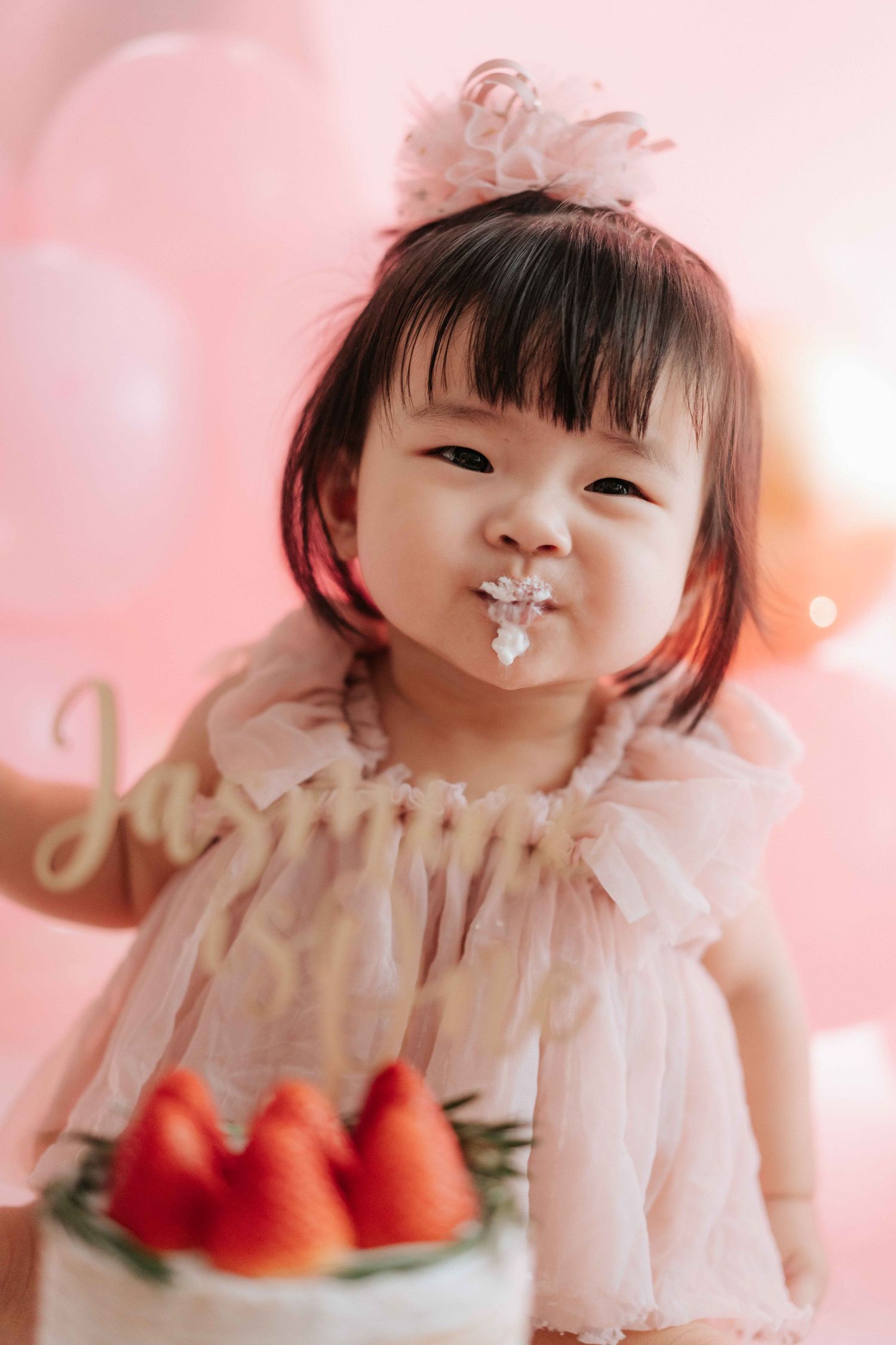 A young child in pink tulle dress poses playfully with strawberries against a soft pink background.