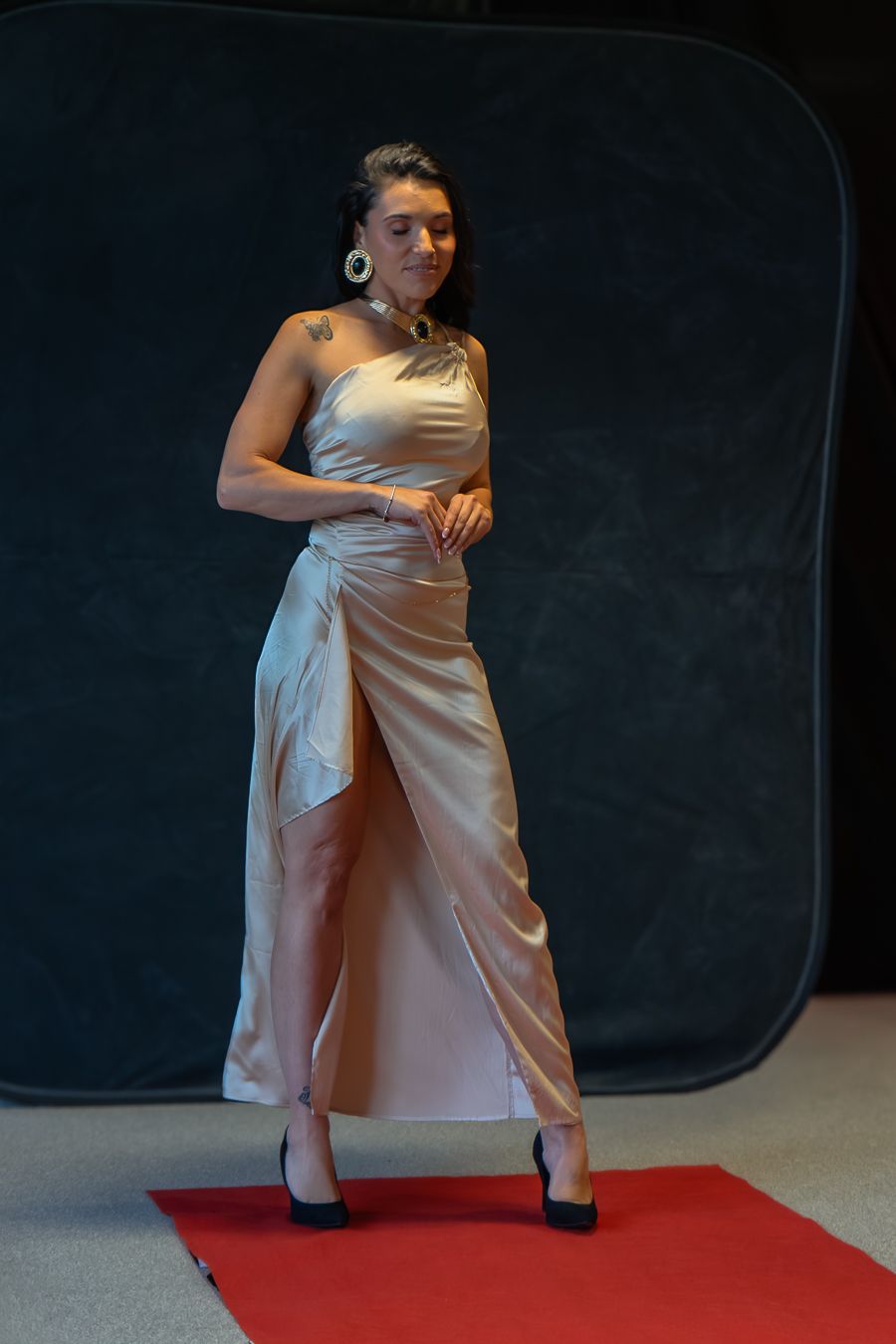 A model poses in a sleek satin evening gown on a red carpet against a dark backdrop.