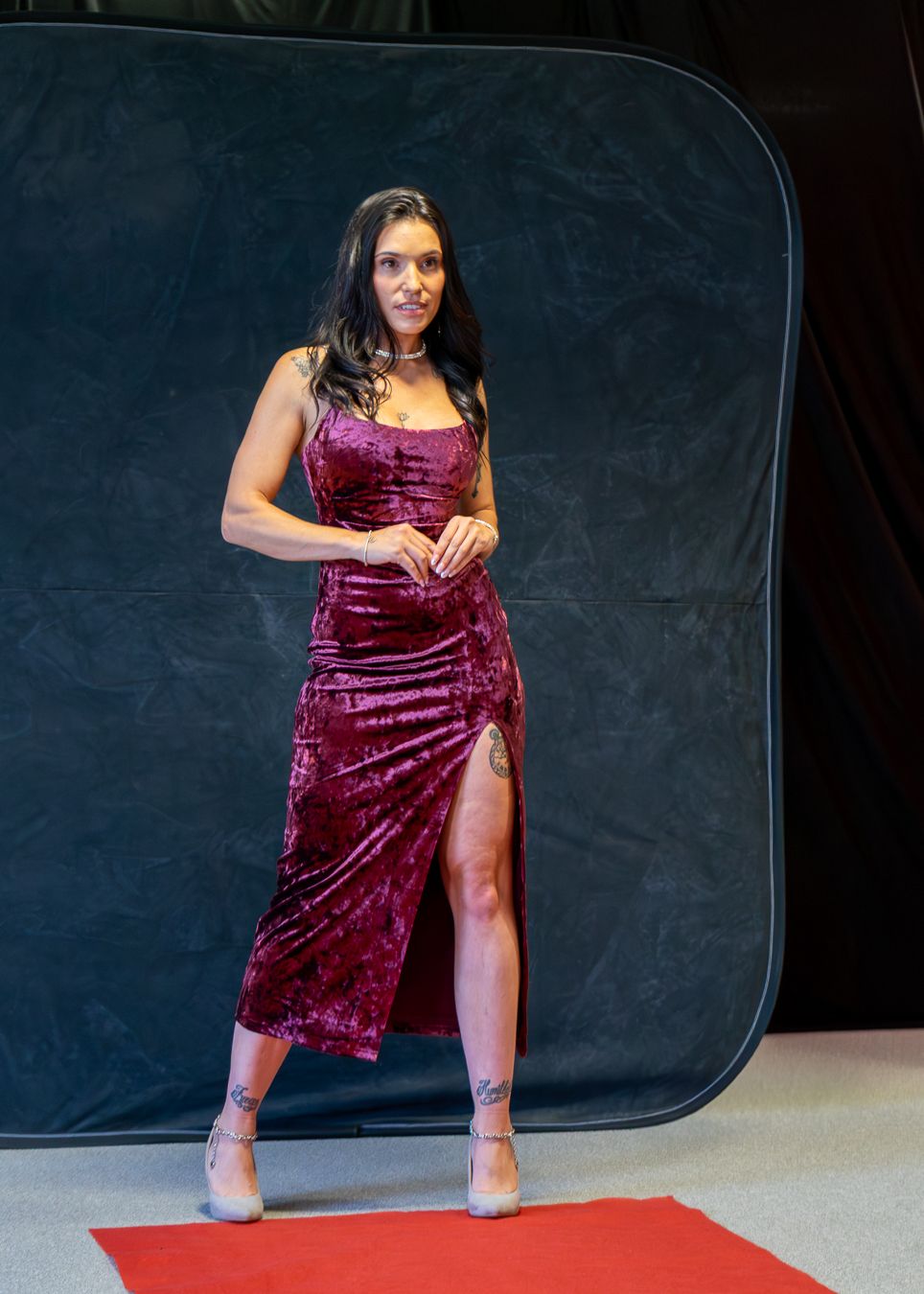 A model poses on a red carpet wearing a burgundy velvet dress with a thigh slit and matching heels against a dark backdrop.