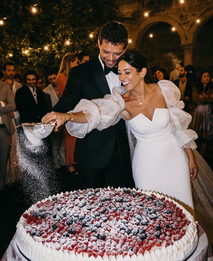 Smiling bride and groom dusting powdered sugar on a large berry wedding cake.