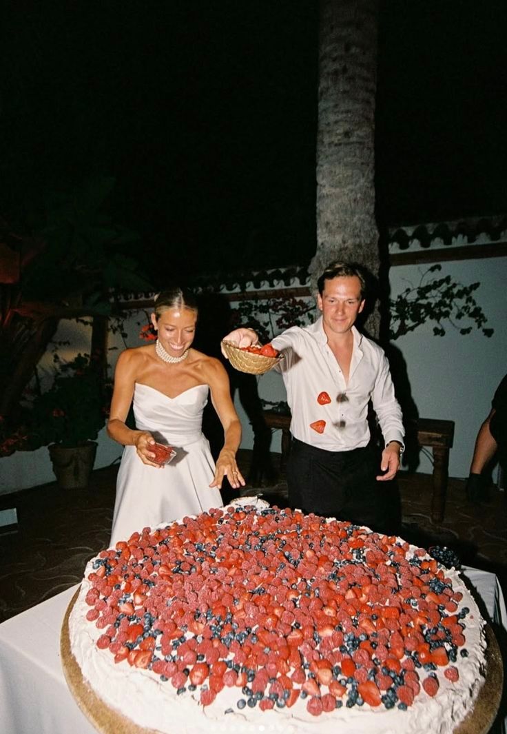Couple cutting a giant berry-topped wedding cake outdoors at a tropical venue.