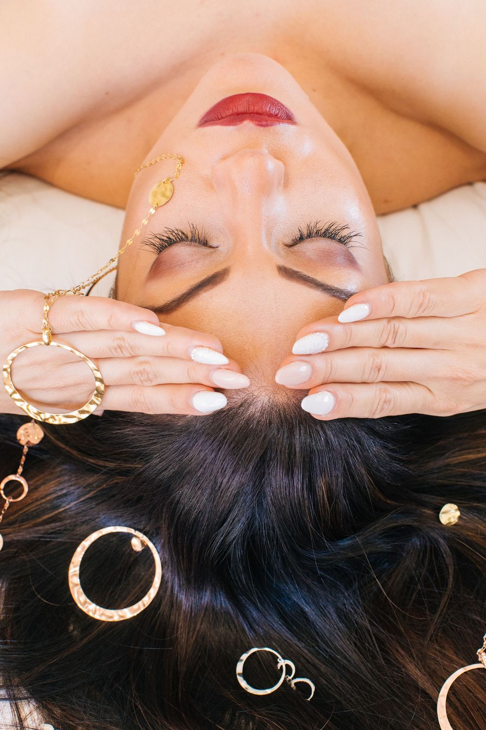 Woman lying down with closed eyes, white nails, red lips, and gold jewelry.
