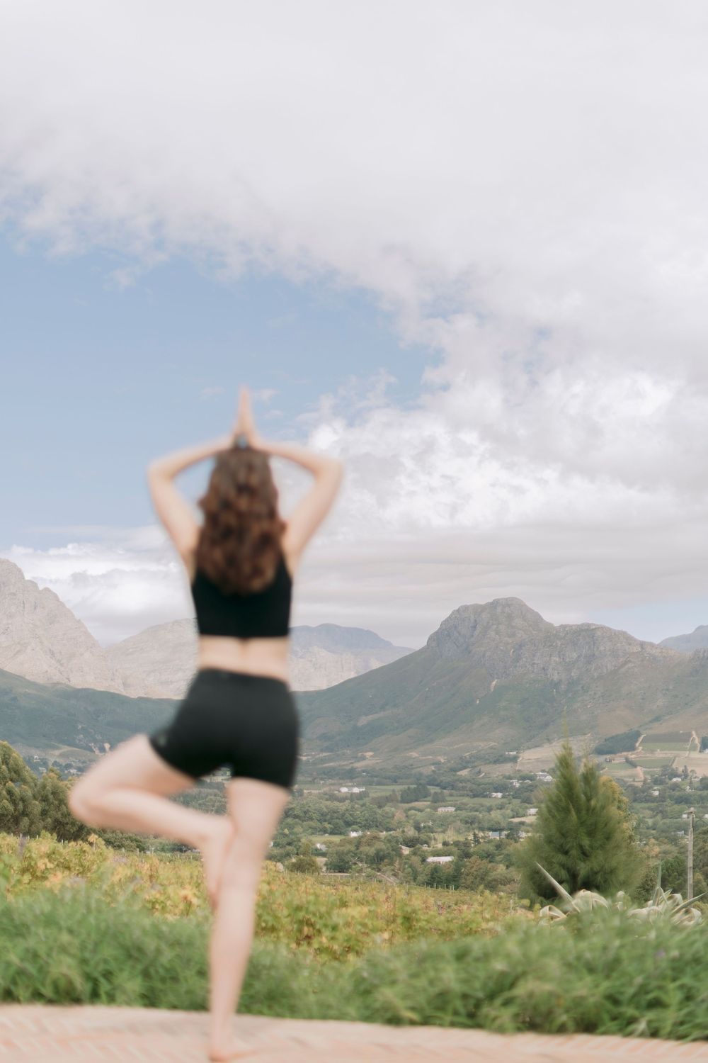 Woman in black activewear practicing tree pose yoga on an outdoor terrace with mountain valley views.