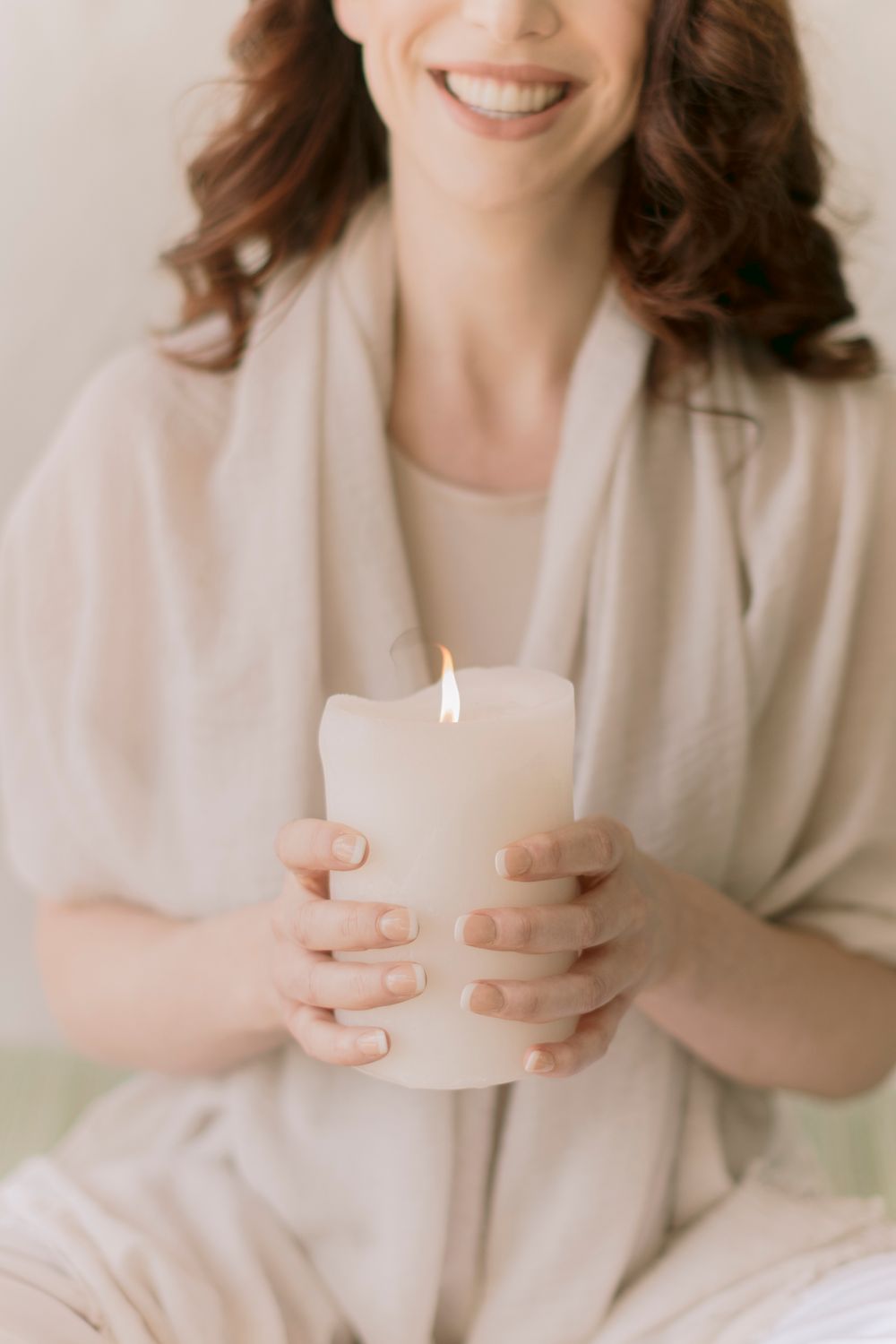 Smiling woman in neutral tones holding a lit white pillar candle while seated indoors.