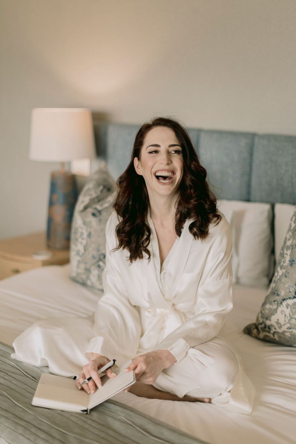 A laughing woman in a white silk robe sits cross-legged on a bed writing in a notebook.