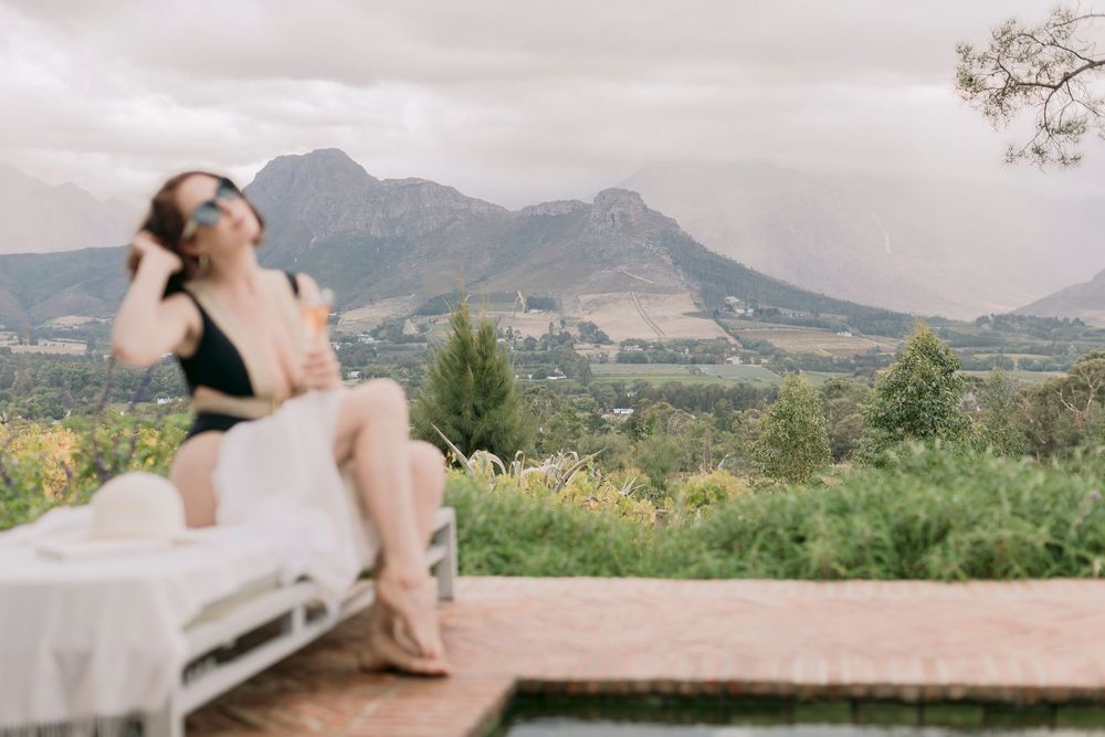 A woman in a black swimsuit sits on a poolside lounger with a stunning mountain backdrop.