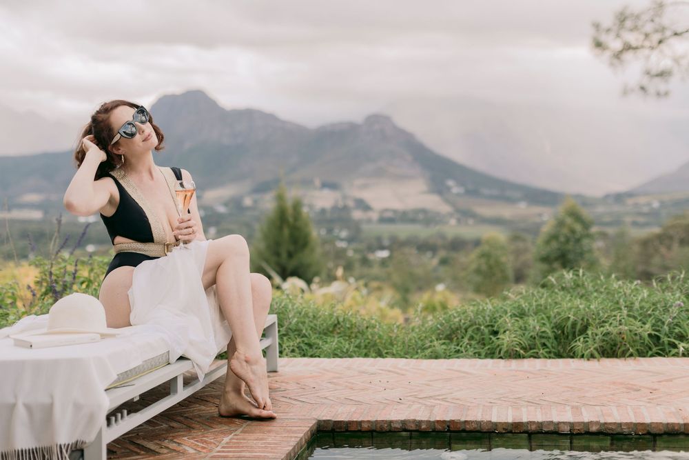 A woman in a black swimsuit and sunglasses enjoys champagne by a pool with mountain views.