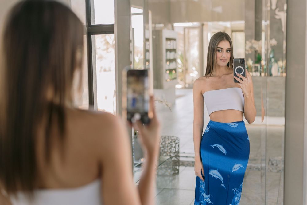Young woman in white crop top and blue skirt taking a mirror selfie in a bright boutique fitting room.