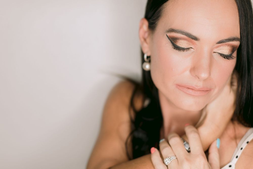 Close-up of a brunette woman with gold eye makeup and pearl earrings, wearing a diamond ring, eyes closed.
