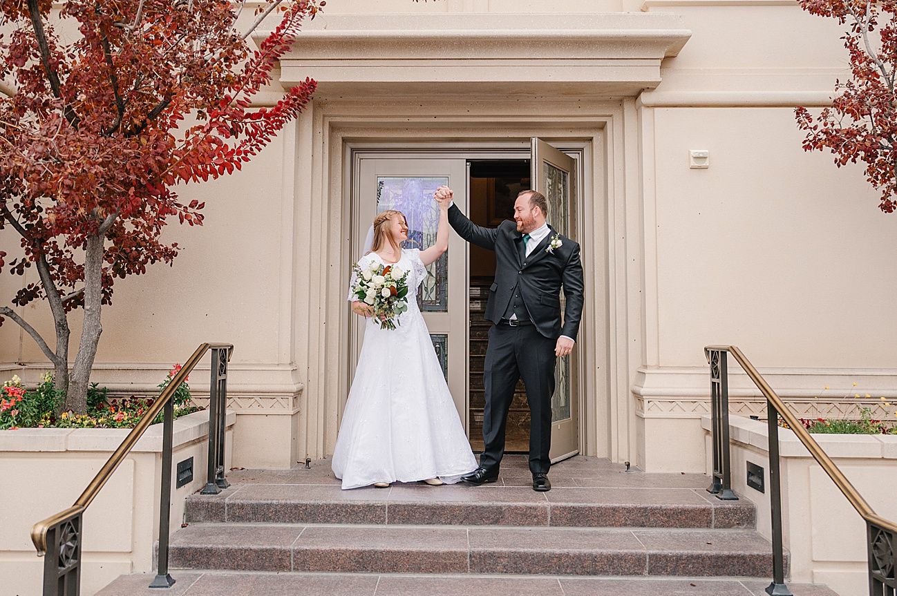 A wedding couple stands in front of a beige building with red autumn trees creating a romantic outdoor scene.
